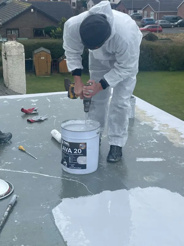 Roofer applying liquid flat roof waterproofing from a bucket with a paint brush, garden visible behind