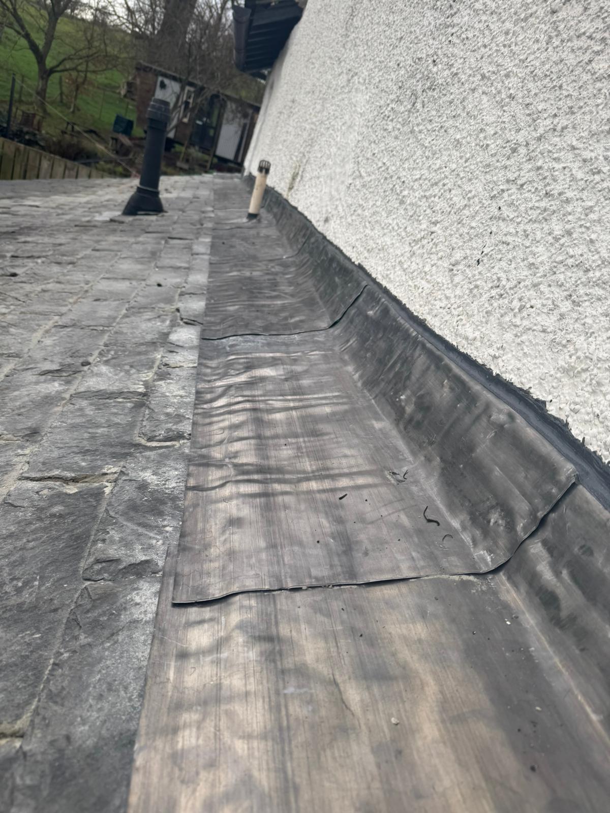 New lead flashing running the full length of an abutment on an old natural slate roof alongside a pebbledash wall, with vent pipes visible